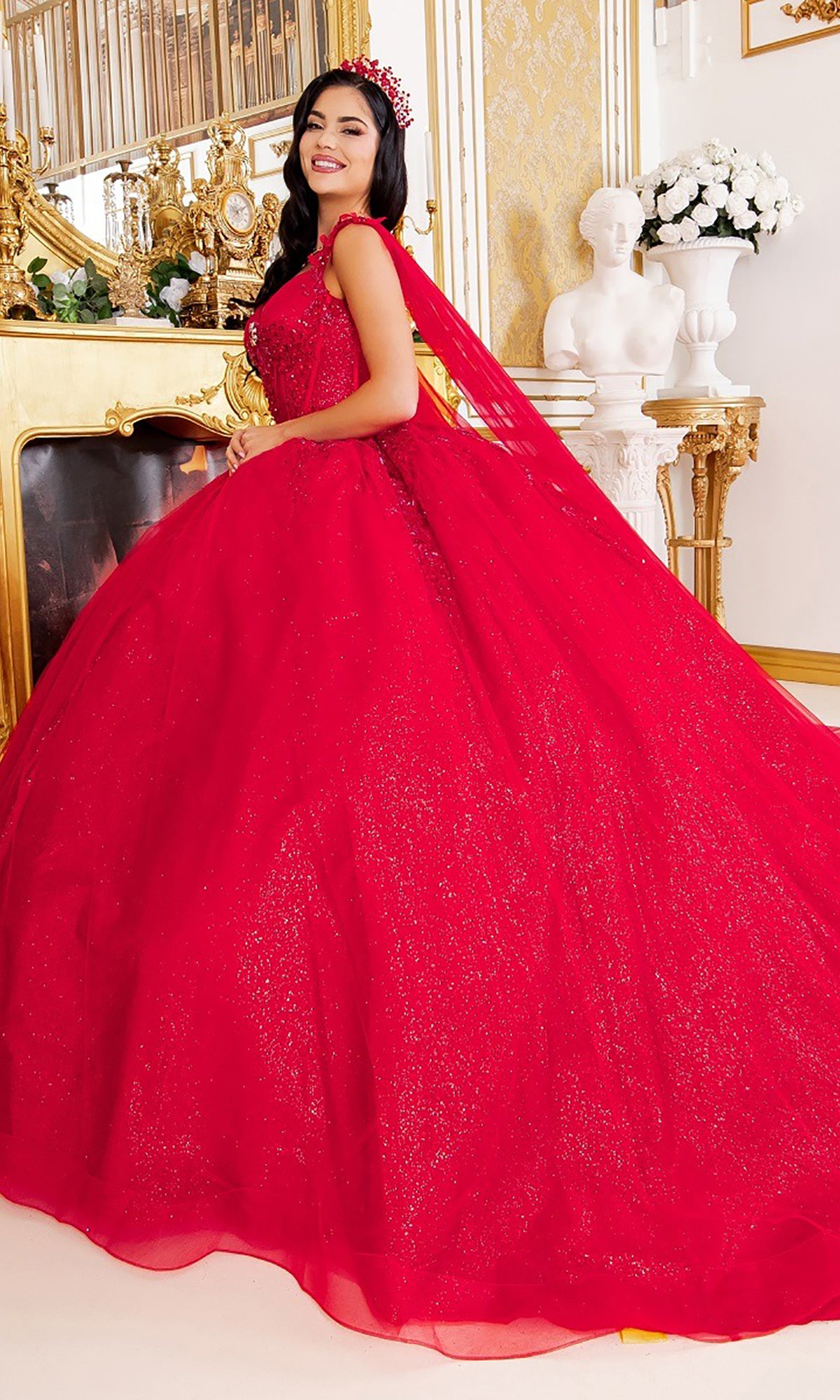 A woman wearing the Quinceanera Dress 8270J By Cinderella Couture—a sparkling red ball gown with elegant cape sleeves and a tiara—stands in an ornate room adorned with gold accents, a fireplace, and a white statue in the background.